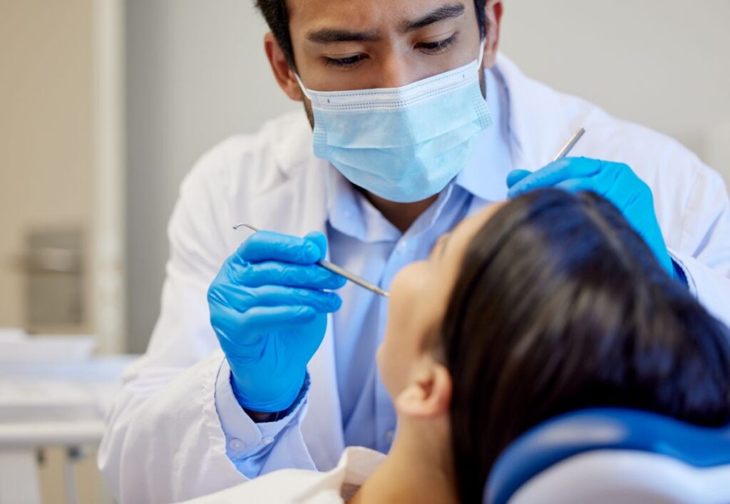 Dentist in a mask and blue gloves performing a dental checkup on a patient in a chair.