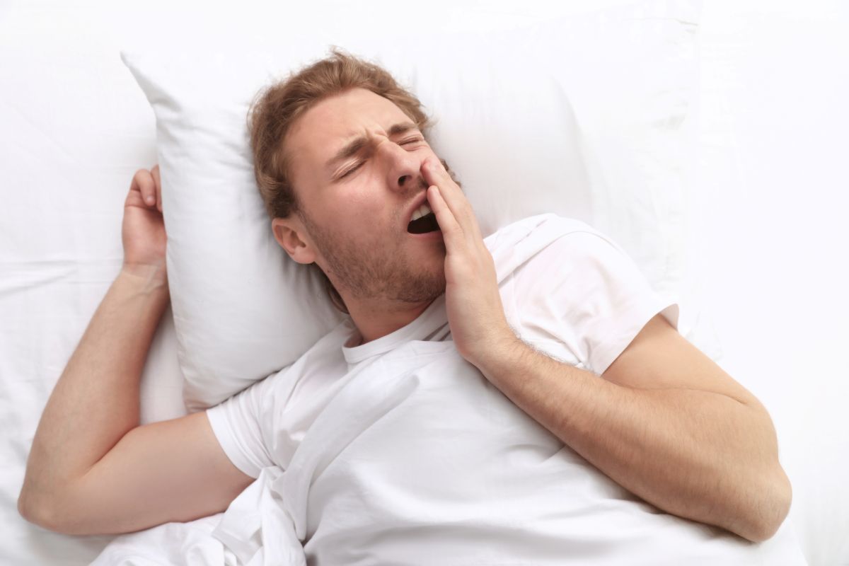 handsome-young-man-yawning-while-lying-on-pillow-i-2026-03-25-23-00-03-utc - Complete Health Dentistry Young man yawning in bed, resting on a white pillow with white sheets.