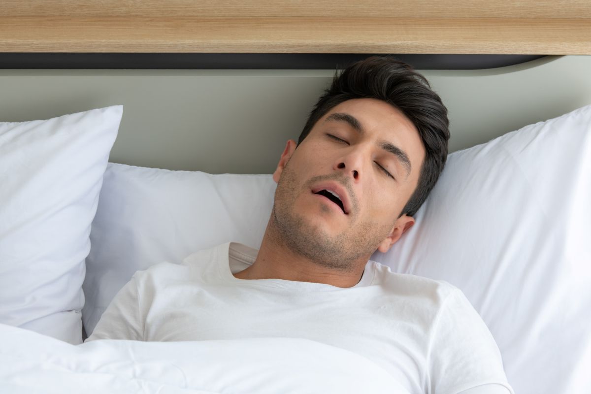 Man sleeping in a white-dued bed with pillows, wearing a white T-shirt and eyes closed under a light blanket.