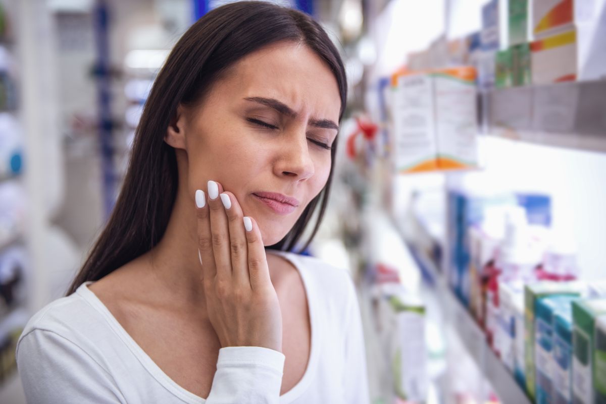Woman in a pharmacy aisle holding her cheek in visible discomfort, suggesting tooth or jaw pain.